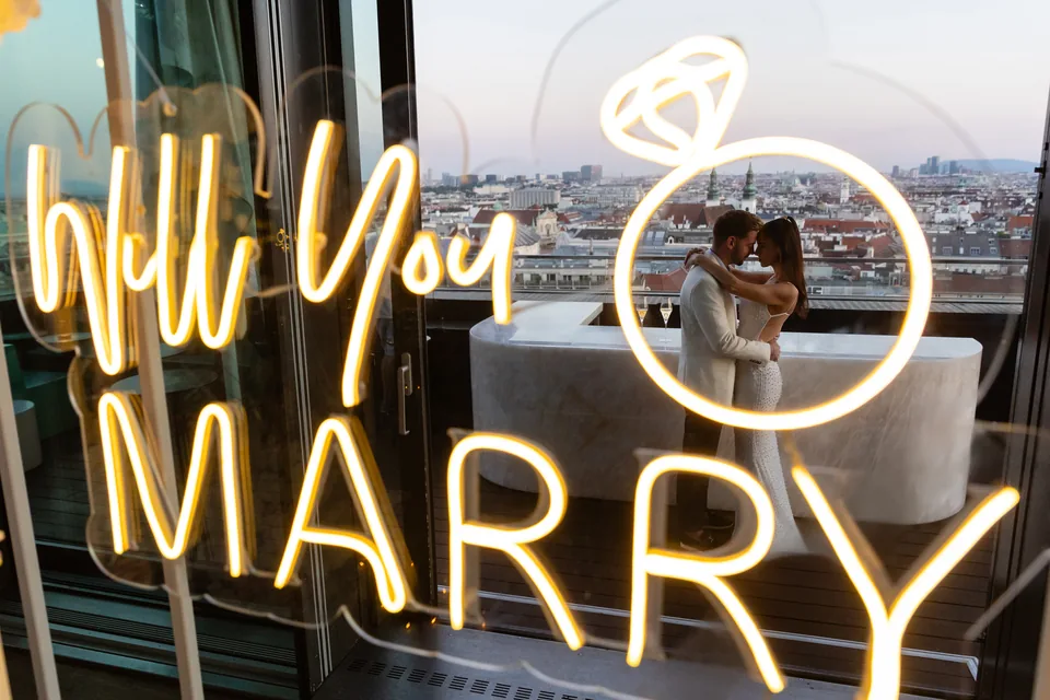 Neon sign reading Will You Marry Me with the newly engaged couple behind it above the rooftops of Vienna